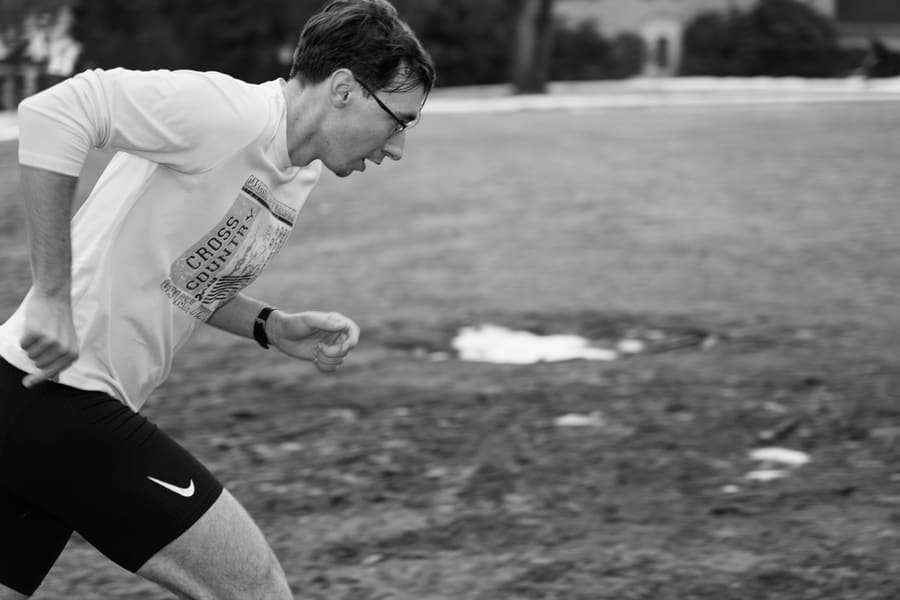 A runner in a cross country shirt sprinting across a field with patches of snow